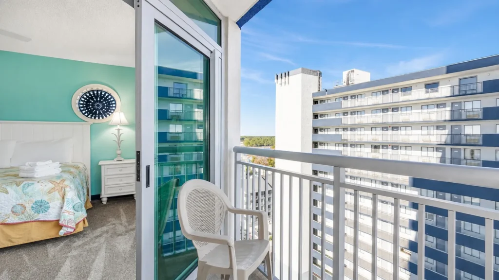Hotel balcony with a white railing and plastic chair, overlooking a blue-sky cityscape and neighboring buildings through a glass door.