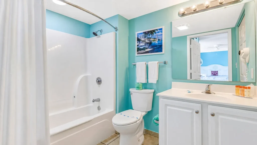 Bright turquoise bathroom with a white tub/shower, toilet, and a white vanity seen from the doorway.