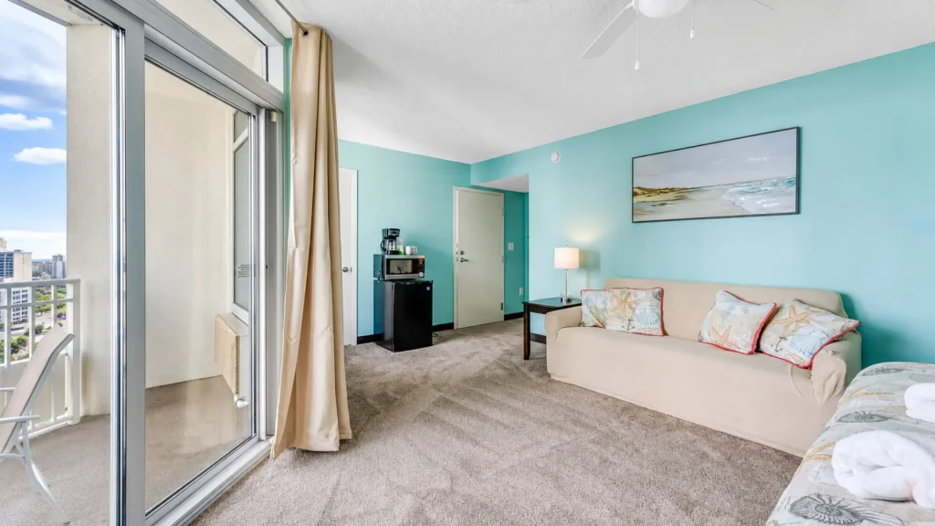 Coastal hotel living area with turquoise walls, beige sofa with sea-shell pillows, and a balcony door to an urban view; kitchenette with microwave nearby.