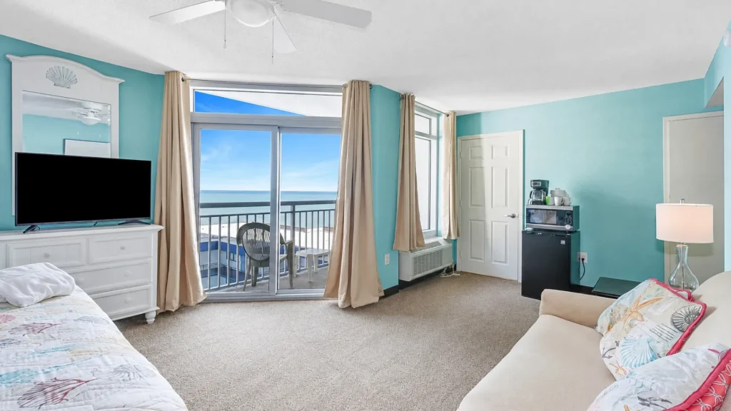 Hotel room with turquoise walls and a sliding glass door to a balcony overlooking the ocean; TV on a white dresser and beige curtains framing the view