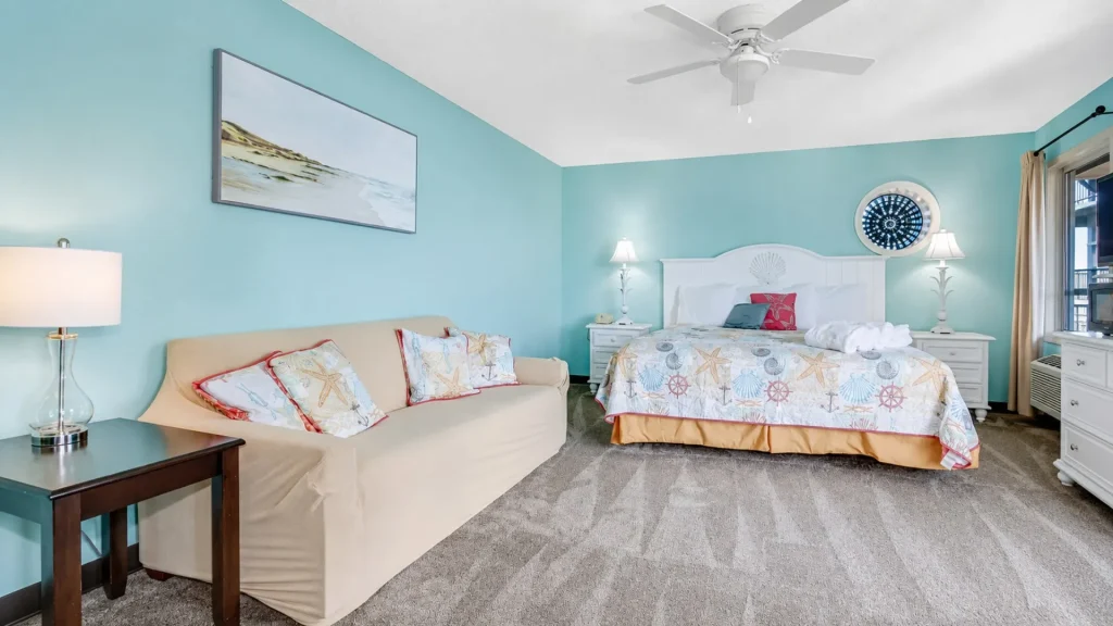 Coastal hotel bedroom with turquoise walls, white bed, seashell quilt, and beige sofa.