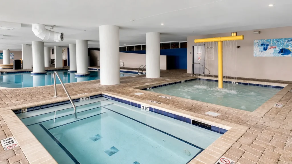 Indoor pool facility with multiple rectangular pools, tile edges, and white support pillars in a spacious room like a spa or gym. A yellow overhead water feature jets water into the rightmost pool.