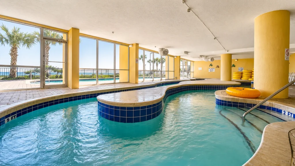 Indoor pool area with large glass windows, yellow columns, and a view of an outdoor pool and palm trees by the sea outside.