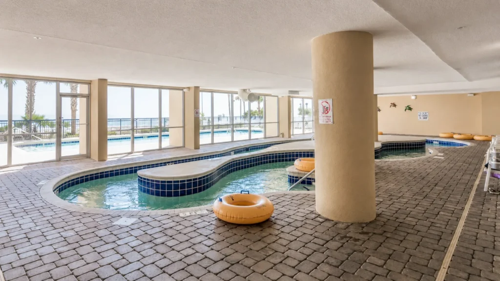 Indoor pool area with curved blue-tiled edge, orange life rings, and a large round pillar, backed by floor-to-ceiling windows showing palm trees and the ocean outside