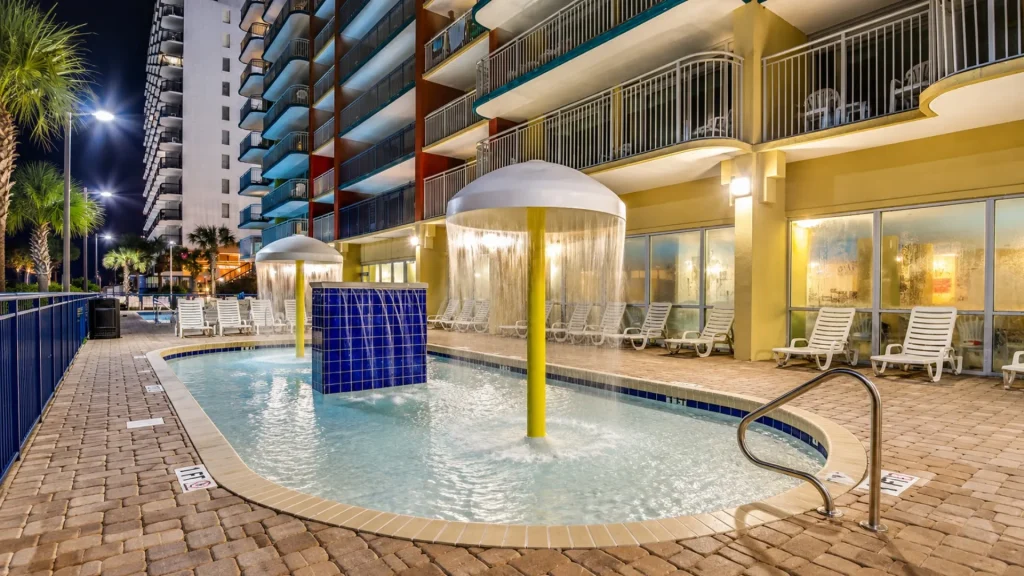 Nighttime hotel pool area with a circular fountain and cascading water, surrounded by lounge chairs and glass-walled hotel rooms.