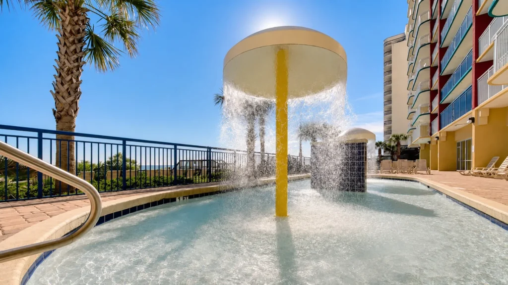 Pool with a bright yellow water feature spraying into the shallow area, palm trees, and a multi-story hotel with balconies on the right under a clear blue sky.
