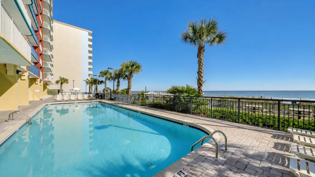 Pool area at a beachfront hotel with blue water, lounge chairs, and a railing overlooking the ocean under a clear sky.