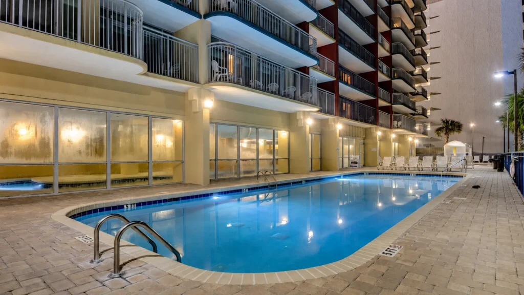 Outdoor hotel pool at night, lined with lounge chairs and adjacent glass-walled amenities.