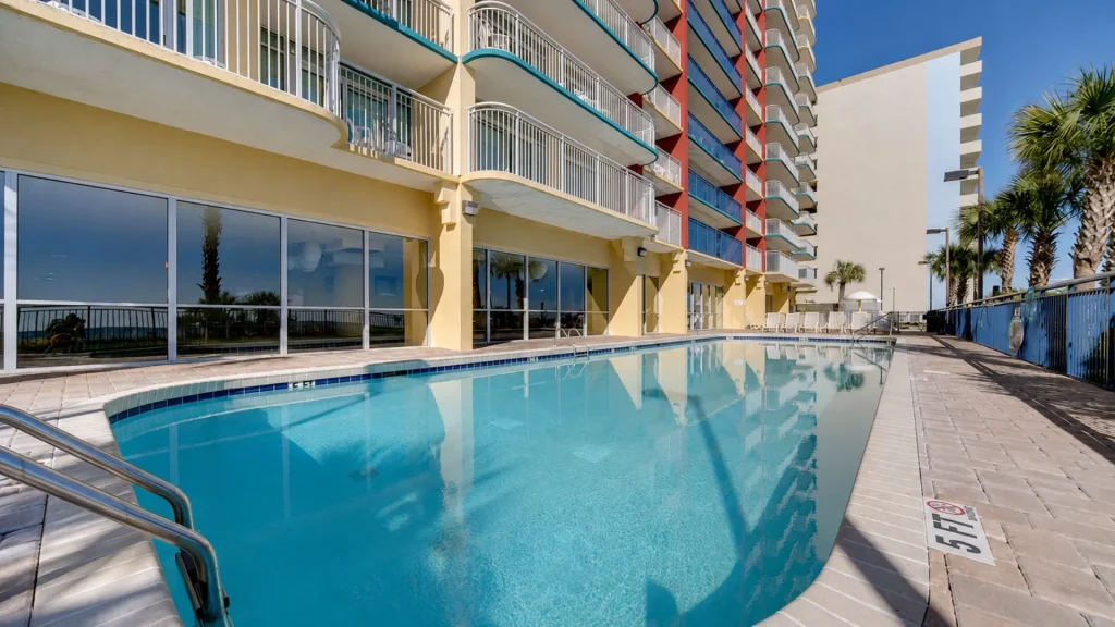 Hotel-style pool area beside a yellow building with colorful balconies and glass walls, palm trees in the background