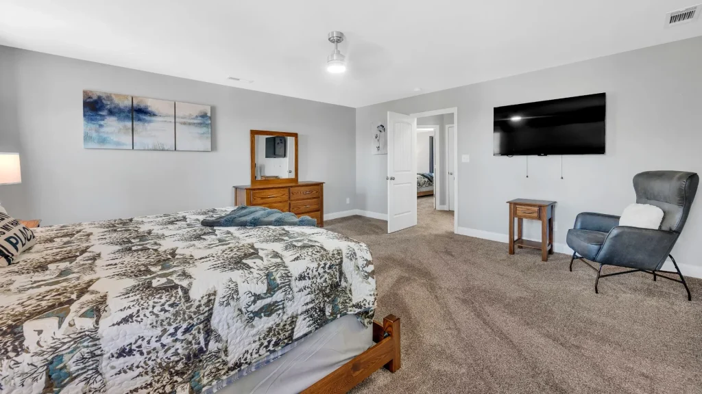 Bedroom with a wooden bed, patterned quilt, dresser with mirror, wall-mounted TV, and a gray recliner chair placed on carpeted floor.