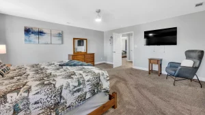 whipering-winds-slider-images 12 | Swack Vacations Bedroom with a wooden bed, patterned quilt, dresser with mirror, wall-mounted TV, and a gray recliner chair placed on carpeted floor.