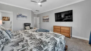 whipering-winds-slider-images 13 | Swack Vacations Bedroom with a patterned comforter, wooden dresser, and a wall-mounted TV; gray walls, ceiling fan, and a black leather chair in the corner.