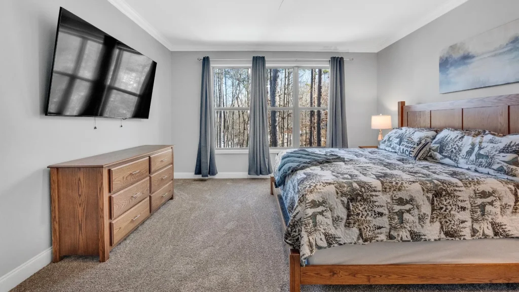 Bedroom with a wooden bed, dresser, wall-mounted TV, and large window with blue curtains; forest outside.