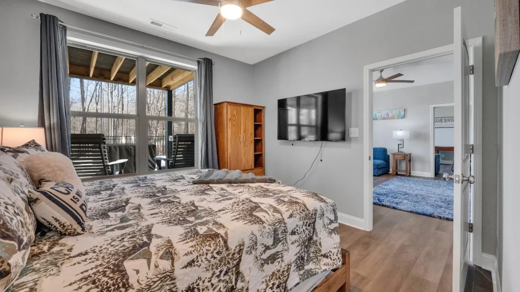 Bedroom with gray walls, large windows to a deck, and a bed with forest-pattern bedding under a ceiling fan