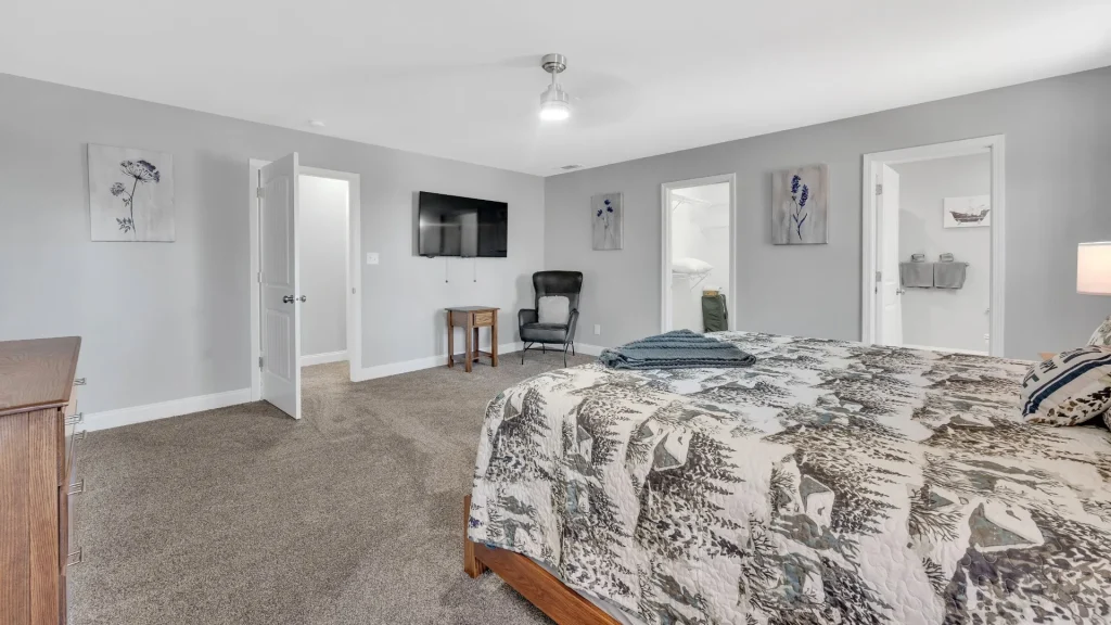 Bedroom with gray walls and a large bed in the foreground, forest-pattern quilt, and a small wooden side table; a wall-mounted flat-screen TV and a black recliner along the far wall.