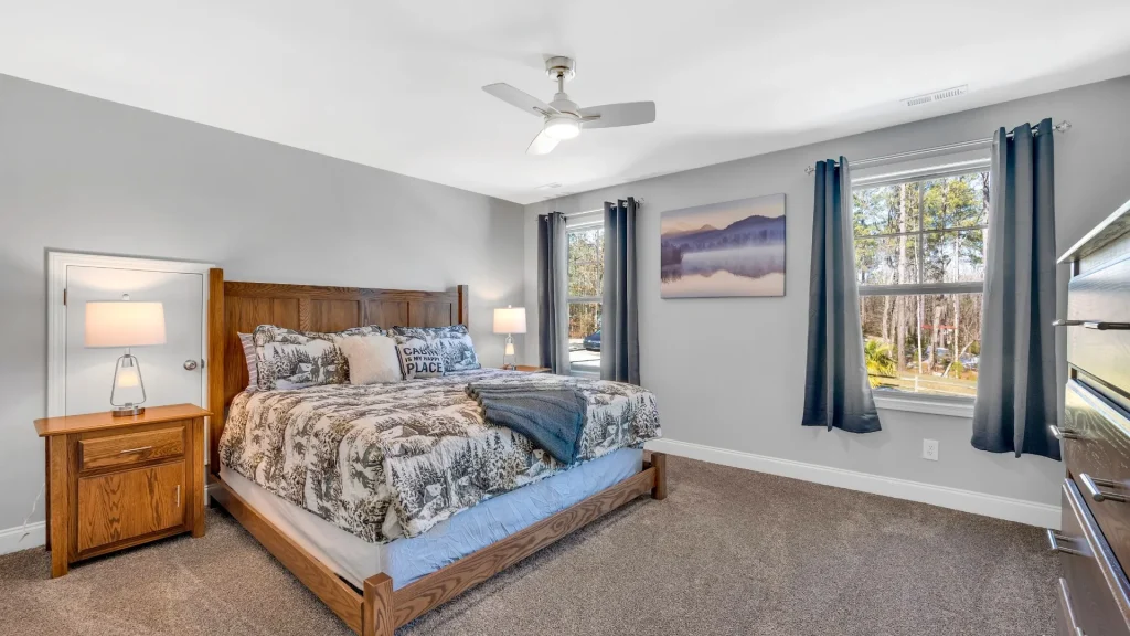 Bedrooom with wooden bed frame, two nightstands, neutral gray walls, and two windows with blue curtains.