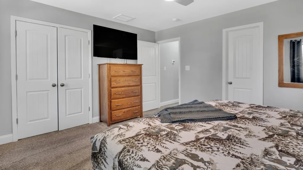 Bedroom with grey walls, a wooden dresser with a TV on top, and white paneled closet doors on the left; bed with patterned bedding in foreground.