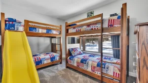 whipering-winds-slider-images 23 | Swack Vacations Two wooden bunk beds with red‑white‑blue patchwork quilts; a yellow slide in the foreground and a snowy window view.