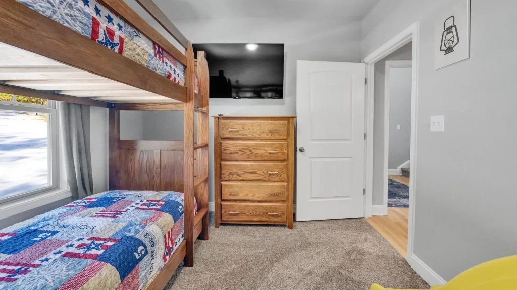 Bedroom with a wooden bunk bed, patchwork quilt, window with gray curtains, and a tall dresser beneath a wall-mounted TV.