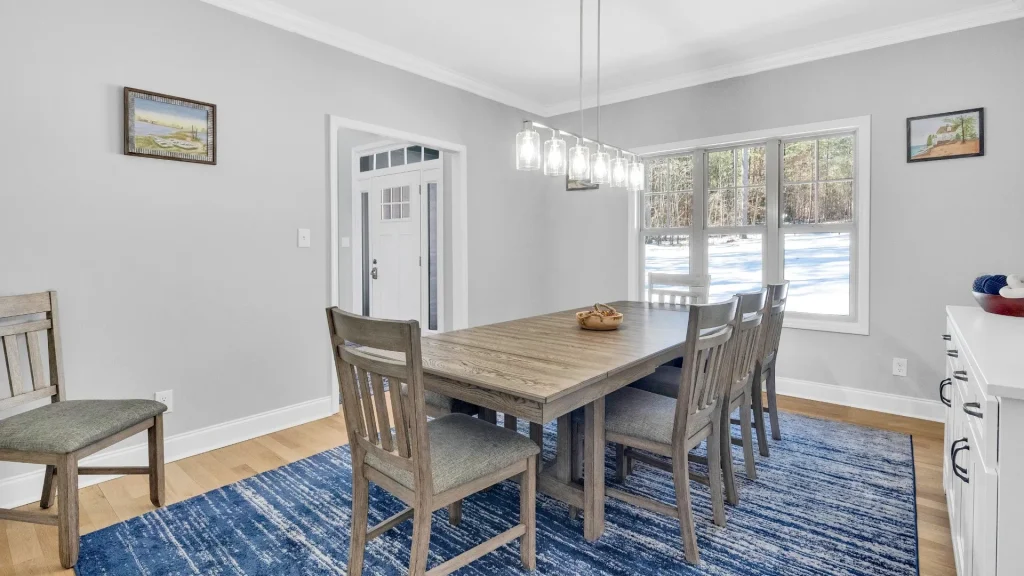 Dining room with a rustic wooden table and six chairs, gray walls, and a blue area rug; large window shows a snowy yard outside.