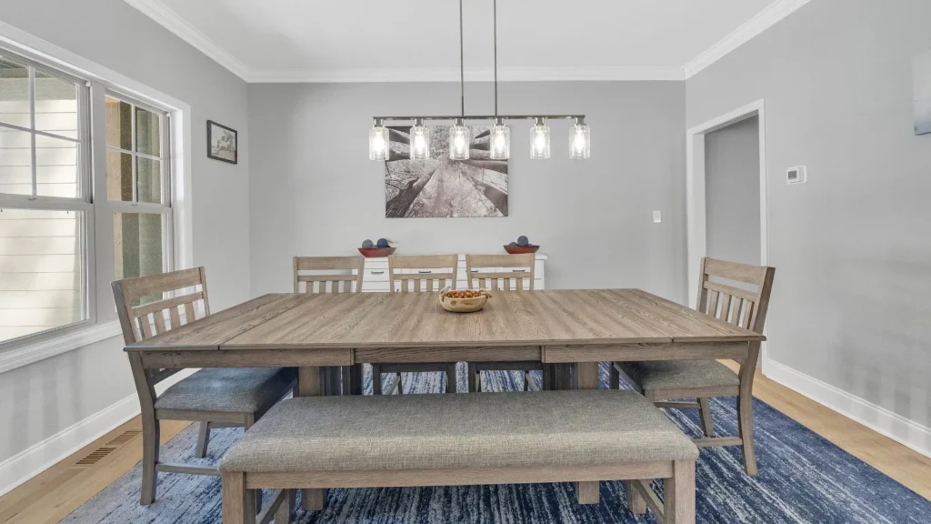 Wood dining table with bench seating and six chairs in a gray dining room, pendant lights overhead and a blue rug underneath.
