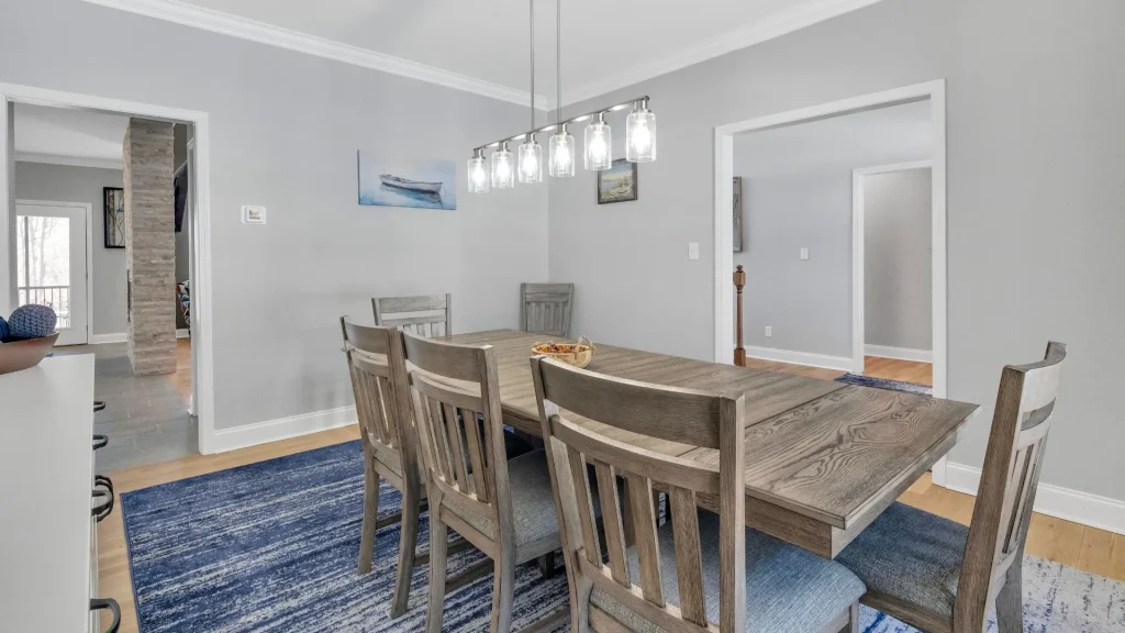 Dining room with a large wooden table and six chairs on a blue rug, gray walls, and a row of glass pendant lights above the table.