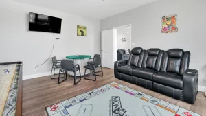 whipering-winds-slider-images 29 | Swack Vacations Living room with a black leather sectional sofa, wall-mounted flat-screen TV, and a round green game table against light gray walls.