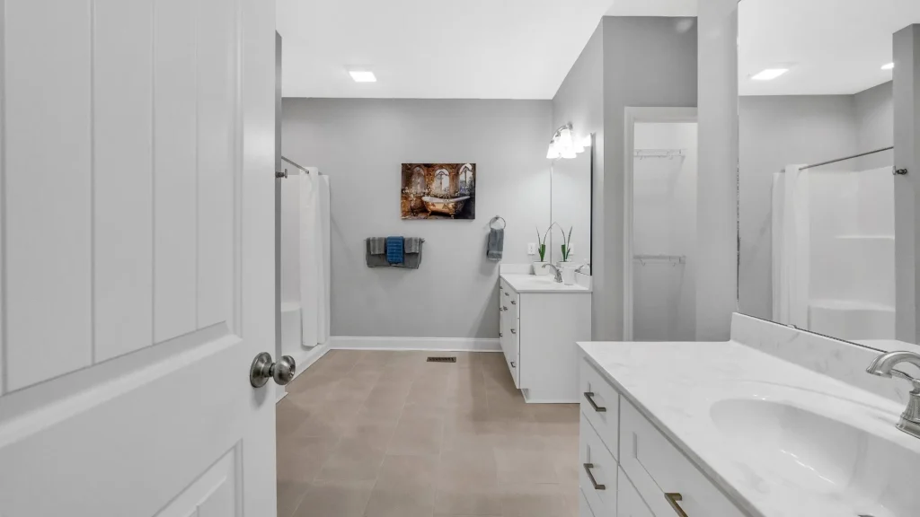 Modern bathroom with a double vanity, gray walls, and a wall art piece; towels on a rack and a shower to the left.