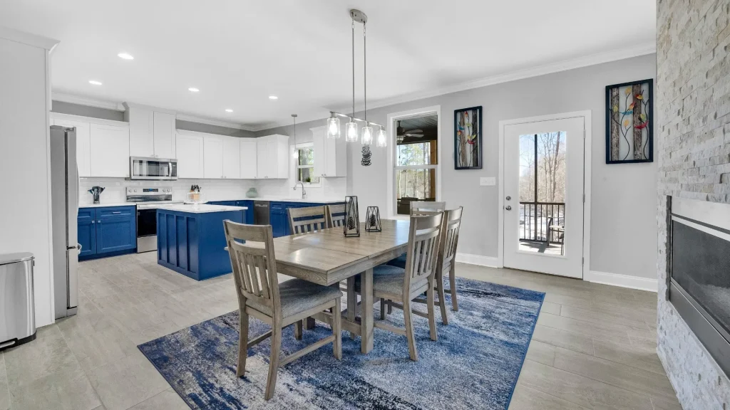 Open-concept kitchen and dining area with blue lower cabinets, white upper cabinets, stainless steel appliances, and a wooden dining table on a blue rug.