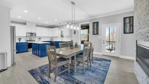 Open-concept kitchen and dining area with blue lower cabinets, white upper cabinets, stainless steel appliances, and a wooden dining table on a blue rug.