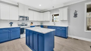 Modern kitchen with white upper cabinets, blue lower cabinets, marble island, stainless appliances, and a stacked-cup wall decal on the gray wall.