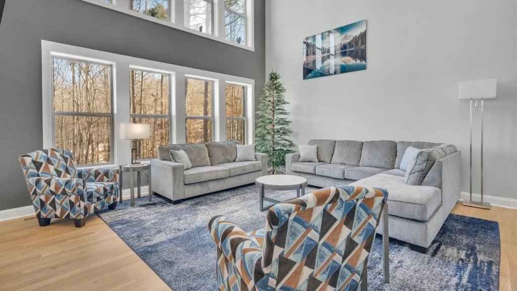 Bright living room with gray sectional sofa, blue patterned armchairs, and a blue rug under a round coffee table by large windows.