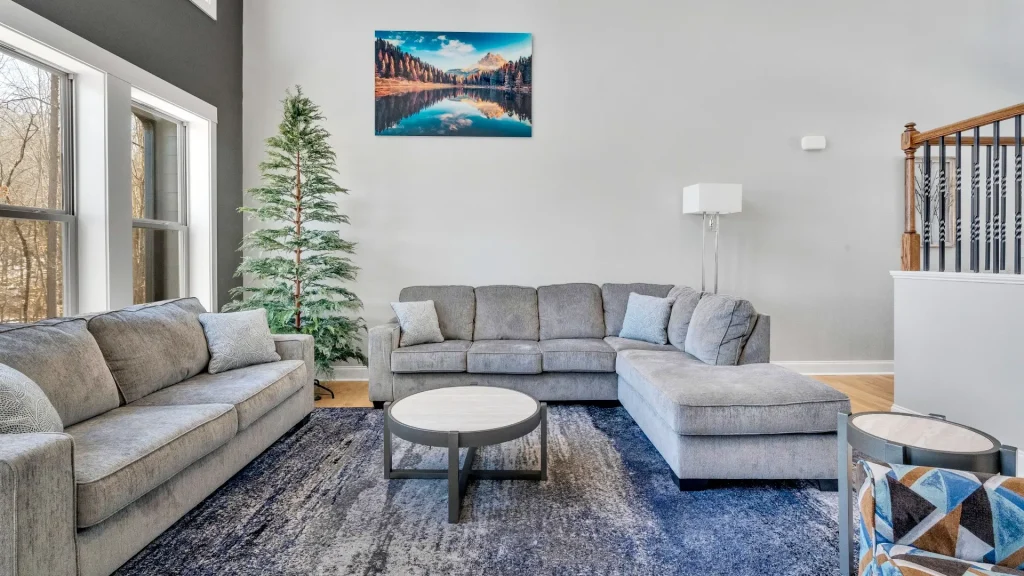 Modern living room with gray sectional sofa, blue-gray rug, and round coffee tables near large windows; a tall plant and landscape wall art in view.