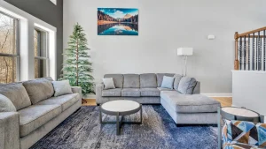 whipering-winds-slider-images 35 | Swack Vacations Modern living room with gray sectional sofa, blue-gray rug, and round coffee tables near large windows; a tall plant and landscape wall art in view.