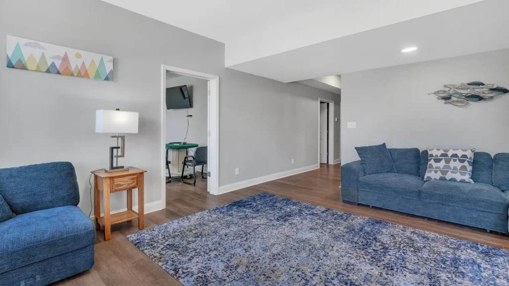 Bright living room with blue sectional sofas, grey walls, and a blue patterned area rug on wood floors.