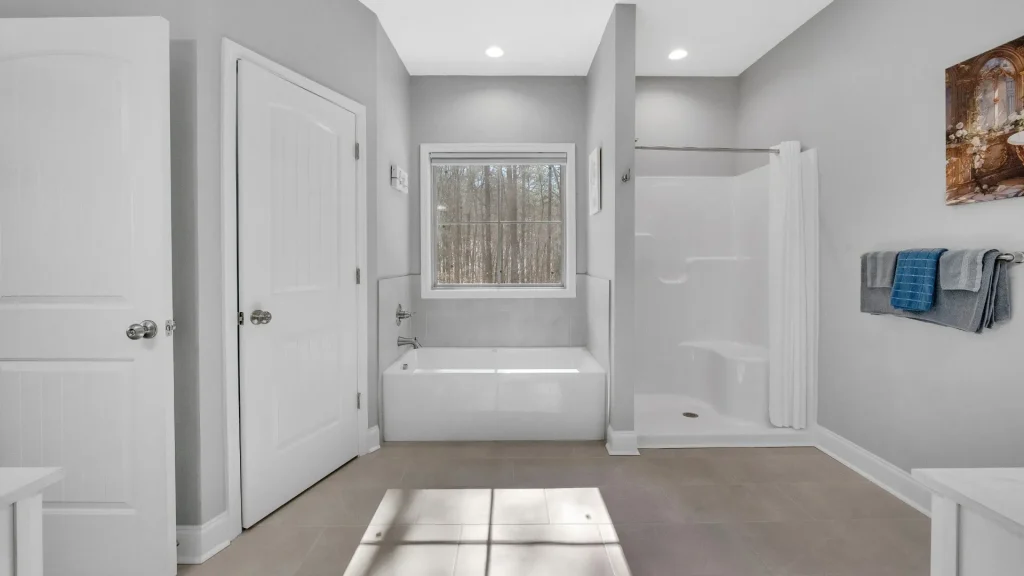 Bright bathroom with a white tub under a window, and a separate shower stall to the right; towels hang on a rack above.