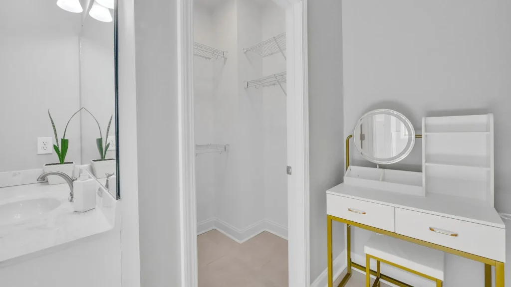 White bathroom with a vanity and two small potted plants, doorway to an open closet with wire shelves in the center, and a white dressing console with a round gold-framed mirror on the right.