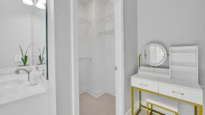 White bathroom with a vanity and two small potted plants, doorway to an open closet with wire shelves in the center, and a white dressing console with a round gold-framed mirror on the right.