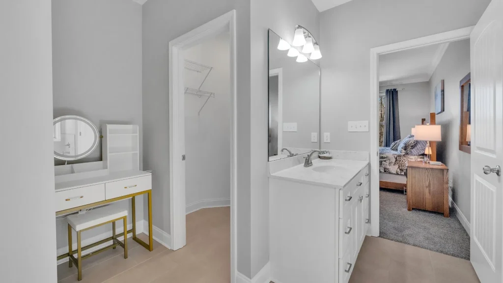 Hallway bathroom with white cabinets and marble countertop, large mirror, and bright light fixtures; doorway opens to a bedroom with a nightstand and bed.