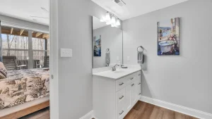 White bathroom vanity with a white countertop, dual faucets, and ample storage beneath; large mirror above, three light fixture, and gray walls with a towel ring and artwork. View into a bedroom area with a bed and sliding glass doors to an outdoor deck.