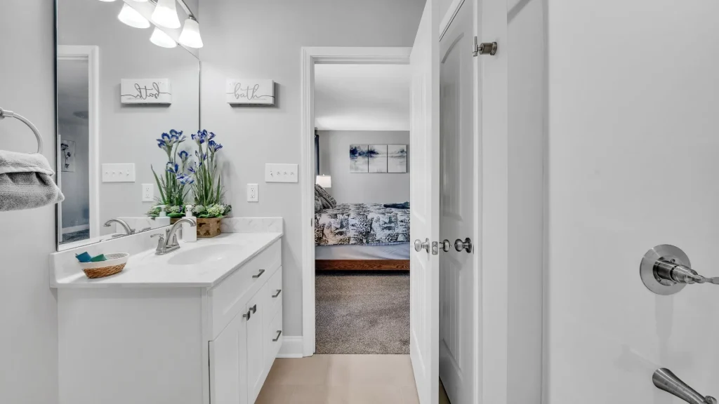 White bathroom vanity with double sinks, potted blue irises, and mirror; open door leads to a bedroom with a bed and art in soft lighting.