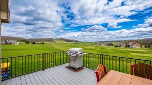 Backyard deck with a stainless grill and wooden table, overlooking a green lawn and distant houses under a cloudy blue sky
