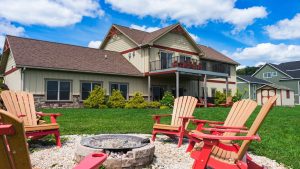 Two-story beige house with red trim and a balcony, surrounded by Adirondack chairs and a circular fire pit in the yard.