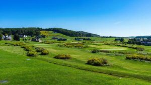 Expansive golf course with rolling greens, shrub beds, and scattered houses under a clear blue sky.