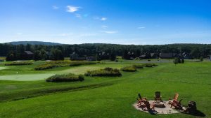 Fire pit with red Adirondack chairs on a grassy hill overlooking a golf course and houses under a clear blue sky.