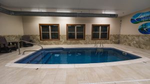 Indoor hotel pool area with three windows along a stone-tile wall and brown lounge chairs on the left side.