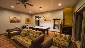 Cozy basement game room with a pool table at center, bear-pattern couches, wood flooring, and wall art including a bear image and a dartboard on the wall.