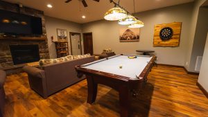 A cozy basement game room with a pool table, sofa, stone fireplace, and dartboard on a wooden wall panel.