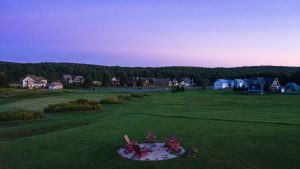 A twilight scene of a suburban neighborhood along a grassy field, with a circular fire pit and red Adirondack chairs in the foreground.