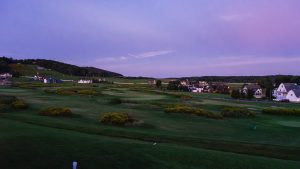 Evening view of a suburban golf course with rolling green fairways and houses along the horizon under a purple sky.